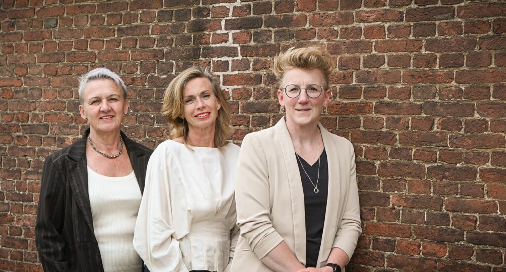 Drie vrouwelijke medewerkers van een rouwdrukkerij staan samen voor een bakstenen muur en kijken vriendelijk in de camera. Rouwkaart katwijk