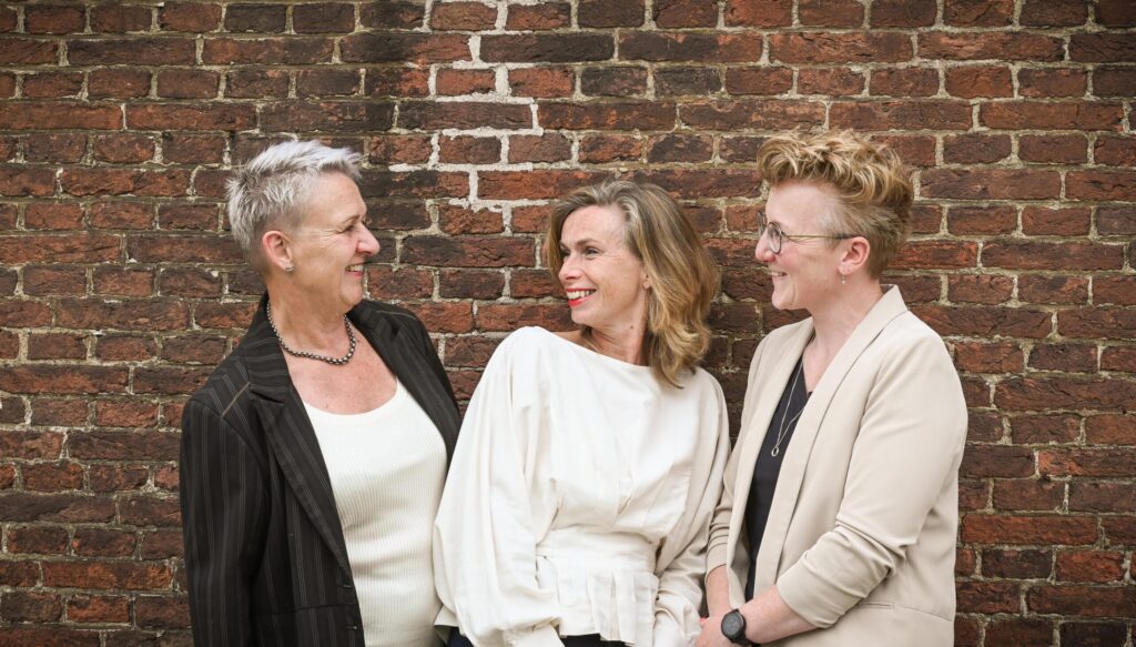 Drie vrouwen staan lachend voor een bakstenen muur – een beeld van betrokkenheid en warmte, met de mogelijkheid om te condoleren: condoleer de familie Katwijk bij overlijdensberichten in Valkenburg.